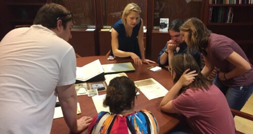 Group of students and instructor examining documents and photographs laid flat on a round table.